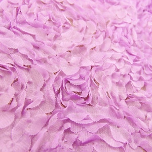 May include: Close-up of a lavender fabric with a floral pattern. The fabric features layered, petal-like designs creating a textured, three-dimensional effect. The delicate, soft material is ideal for crafting or apparel.