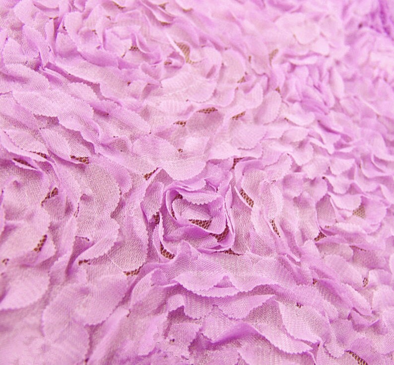 May include: Close-up of a lavender fabric with a floral pattern. The fabric features layered, petal-like designs creating a textured, three-dimensional effect. The delicate, soft material is ideal for crafting or apparel.