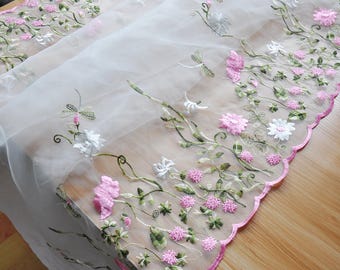 May include: White sheer fabric with pink and white embroidered flowers and green leaves. The fabric has a scalloped edge.