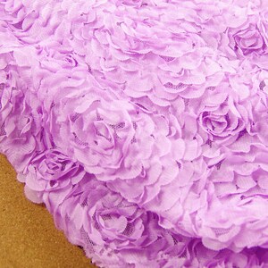 May include: Close-up of a lavender fabric with a 3D floral rose design. The fabric has a delicate, textured appearance, with multiple rose shapes densely arranged across the surface. The color is a soft, pastel purple.