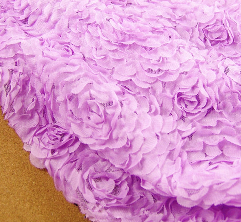 May include: Close-up of a lavender fabric with a 3D floral rose design. The fabric has a delicate, textured appearance, with multiple rose shapes densely arranged across the surface. The color is a soft, pastel purple.