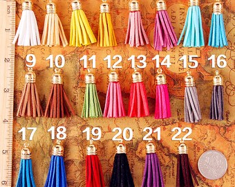 Puede incluir: Una colección de 22 borlas coloridas con tapas doradas, dispuestas sobre un fondo de mapa. Las borlas varían en color, incluyendo blanco, amarillo, rosa, azul y negro. Una regla en pulgadas es visible a la izquierda.