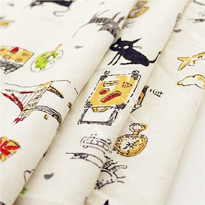 May include: Off-white fabric with a repeating pattern of whimsical illustrations. The design includes black cats, suitcases, clocks, and buildings in shades of yellow, red, and gray. The fabric is folded, showing the pattern's repeating nature.