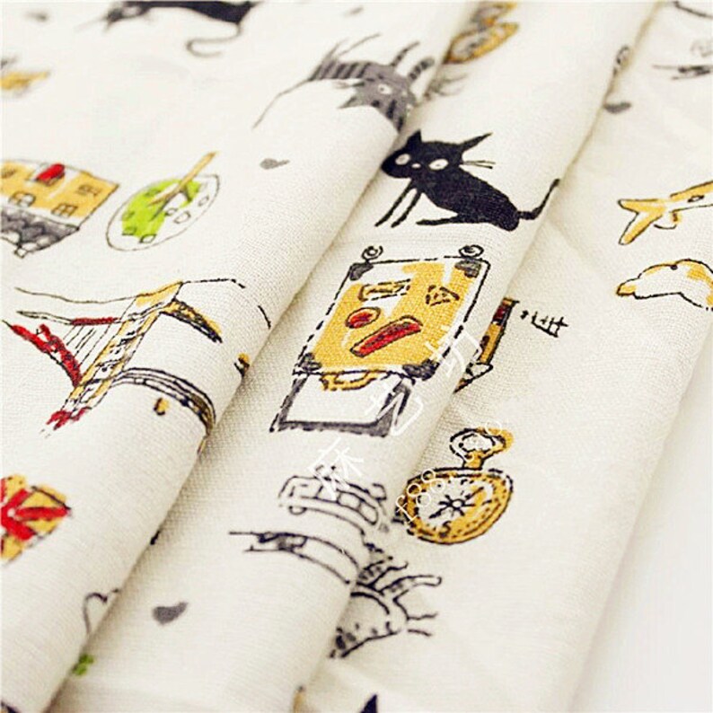 May include: Off-white fabric with a repeating pattern of whimsical illustrations. The design includes black cats, suitcases, clocks, and buildings in shades of yellow, red, and gray. The fabric is folded, showing the pattern's repeating nature.