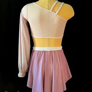 May include: A lilac dance skirt with a white waistband and a matching lilac long-sleeved top with a one-shoulder design and white straps. The top is made of a sheer fabric.