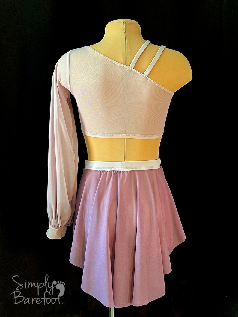 May include: A lilac dance skirt with a white waistband and a matching lilac long-sleeved top with a one-shoulder design and white straps. The top is made of a sheer fabric.