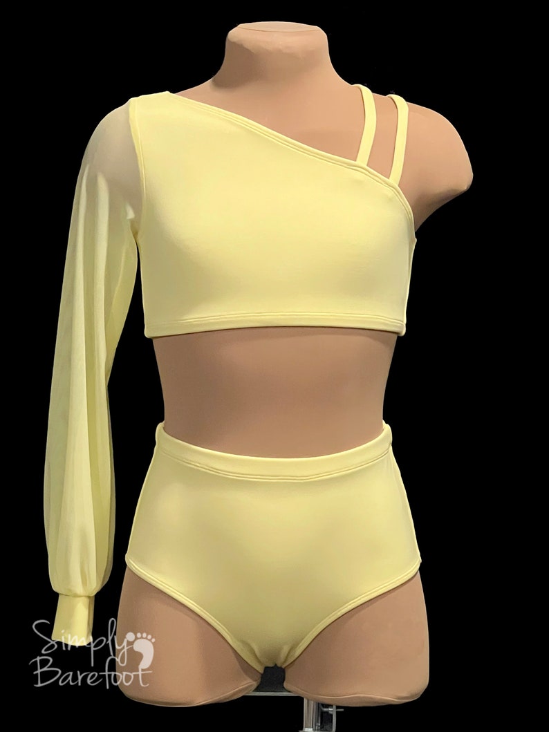 May include: A yellow dance outfit with a one-shoulder crop top and high-waisted shorts. The top has a long sheer sleeve on one side and three straps on the other shoulder. The shorts have a high waist and a fitted silhouette.