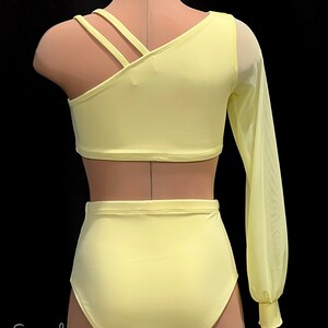May include: A yellow dance leotard with a one-shoulder design and a long sheer sleeve. The leotard has a criss-cross strap detail on the shoulder. The leotard is paired with a matching yellow high-waisted dance skirt.