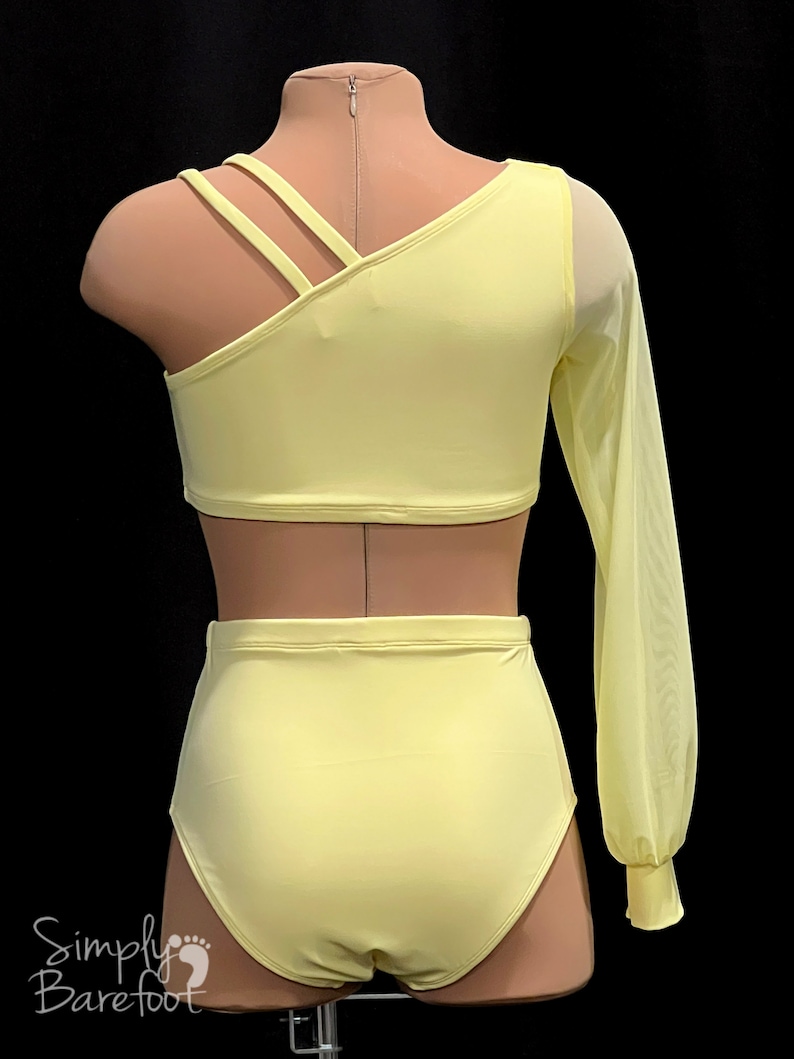 May include: A yellow dance leotard with a one-shoulder design and a long sheer sleeve. The leotard has a criss-cross strap detail on the shoulder. The leotard is paired with a matching yellow high-waisted dance skirt.