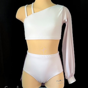 May include: A two-piece dance costume in a light pink color. The top is a one-shoulder crop top with a single long sleeve on the right side. The bottom is a high-waisted skirt with a ruffle at the bottom. The costume has the text "IMINERA 75/59/77/128" printed on the bottom.