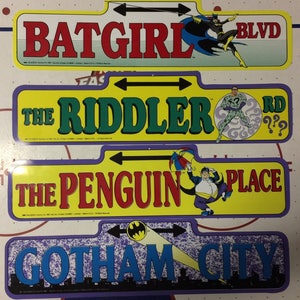 May include: Four colorful street signs with arrows pointing left and right. The signs feature the names of fictional locations from the Batman comics: Batgirl Blvd, The Riddler Rd, The Penguin Place, and Gotham City.