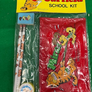 May include: A red pencil case with a zipper closure features a cartoon image of Garfield the cat hanging from a rope. The case comes with three pencils, a pencil sharpener, and an eraser. The pencils have a Garfield cartoon design. The eraser is a small square with a cartoon image of Garfield.