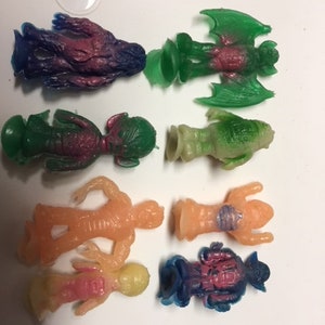 May include: A collection of eight small, colorful, rubber toys. The toys are shaped like aliens, monsters, and robots. They are all different colors, including pink, blue, green, and yellow.