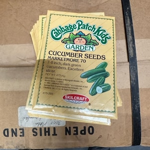 May include: A package of Cabbage Patch Kids Garden Marketmore 70 cucumber seeds. The package is yellow with a green and white illustration of a cucumber. The text on the package reads "Cabbage Patch Kids Garden Cucumber Seeds Marketmore 70 7-8 inch, dark green cucumbers. Excellent slicer. Net Wt. 475 mg Skilcraft".