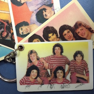 May include: A vintage keychain featuring a group of young men in striped shirts, with a metal chain. Several retro-style cards with similar images are scattered around the keychain. The images have a yellow and white background.