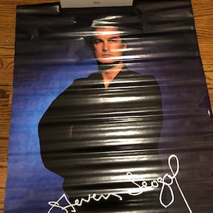 May include: A black and white poster featuring a man wearing a black shirt and a blue background. The man's name is written in white below him: "Steven Seagal".