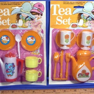 May include: Two vintage plastic tea sets for children, each with a teapot, cups, saucers, a spoon, and a sugar bowl. The teapots are orange and yellow with floral designs. The cups and saucers are yellow and white with floral designs. The sets are packaged on a pink and white background with the words "Tea Set" in large, bold letters.