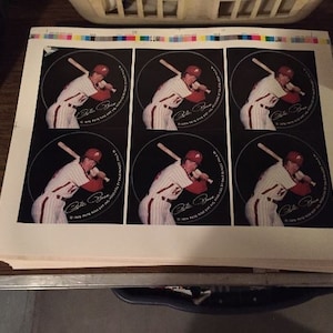 May include: A sheet of baseball stickers featuring Pete Rose in a batting stance. Each sticker is a circle with a black background and the player's image. The stickers are printed on a white sheet with color calibration bars at the top.