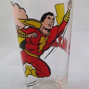 May include: A clear glass with a red and yellow illustration of Captain Marvel, a superhero, flying with a lightning bolt. The glass has the words "Captain Marvel" printed on the side.