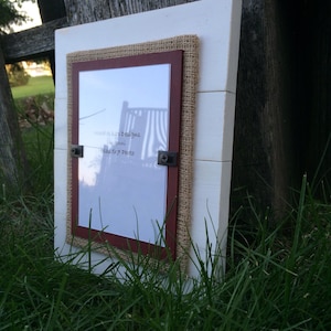 May include: White wooden picture frame with a burlap and red mat. The frame is designed to hold a 5x7 photo. The text "Wood N' Knot Designs" is written on the mat.