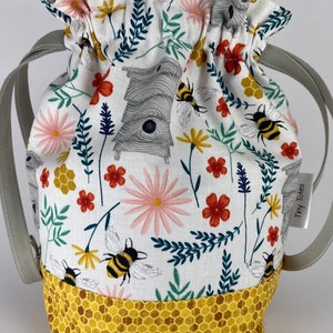 May include: A white drawstring bag with a bee and flower pattern. The bag has a honeycomb pattern on the bottom and two gray straps. The bag has a label that says "Tiny Totes".