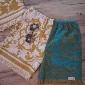 May include: Two rectangular terry cloth towels with fringed edges and a floral pattern. One towel is white with mustard yellow patterns, and the other is teal with mustard yellow patterns. A pair of dark-tinted sunglasses rests on the white towel.