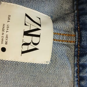 K&ouml;nnte beinhalten: Nahaufnahme eines Jeanskleidungsst&uuml;cks mit einem wei&szlig;en Etikett. Das Etikett tr&auml;gt den Markennamen "ZARA" in fetten schwarzen Buchstaben. Das Kleidungsst&uuml;ck ist aus blauem Denim mit sichtbaren N&auml;hten.