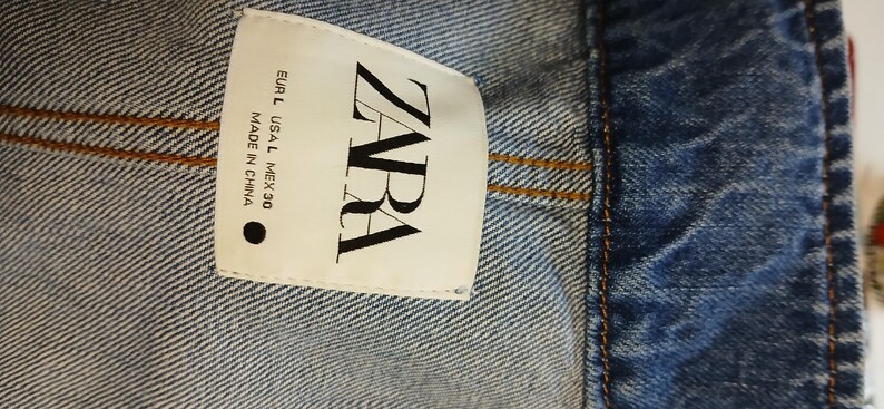 K&ouml;nnte beinhalten: Nahaufnahme eines Jeanskleidungsst&uuml;cks mit einem wei&szlig;en Etikett. Das Etikett tr&auml;gt den Markennamen "ZARA" in fetten schwarzen Buchstaben. Das Kleidungsst&uuml;ck ist aus blauem Denim mit sichtbaren N&auml;hten.