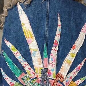 K&ouml;nnte beinhalten: Jeansjacke mit einem farbenfrohen, gesteppten Sonnenmuster auf dem R&uuml;cken. Das Sonnenmuster zeigt verschiedene gemusterte Stoffe in Rosa, Gr&uuml;n und Wei&szlig;, mit einem ausgefransten Saum.