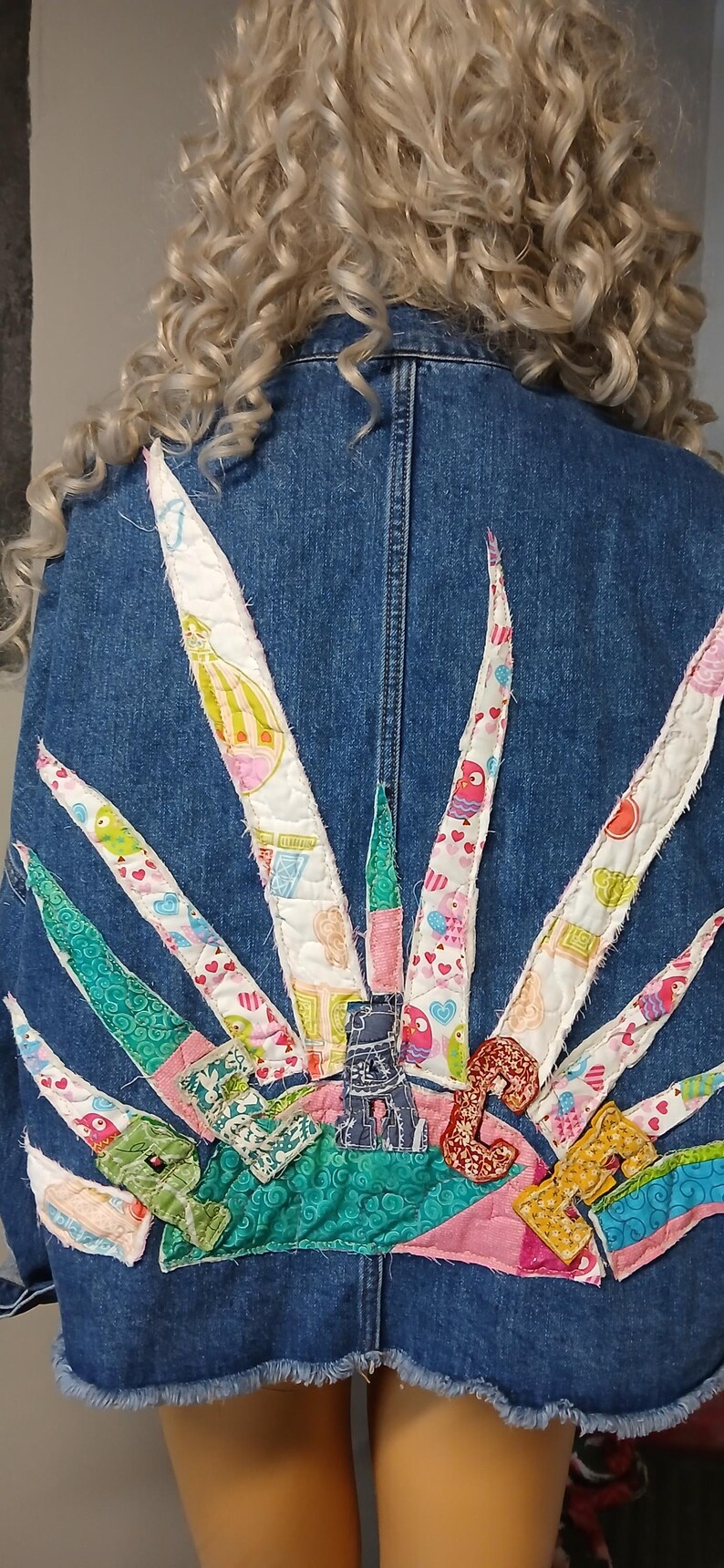 K&ouml;nnte beinhalten: Jeansjacke mit einem farbenfrohen, gesteppten Sonnenmuster auf dem R&uuml;cken. Das Sonnenmuster zeigt verschiedene gemusterte Stoffe in Rosa, Gr&uuml;n und Wei&szlig;, mit einem ausgefransten Saum.