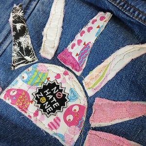 K&ouml;nnte beinhalten: Nahaufnahme eines Jeanskleidungsst&uuml;cks mit bunten Stoffflicken. Zu den Flicken geh&ouml;ren ein schwarz-wei&szlig;es Design, rosa Herzen und Eulen. Ein Aufn&auml;her lautet "NO HATE ZONE" in einem schwarzen Stern.