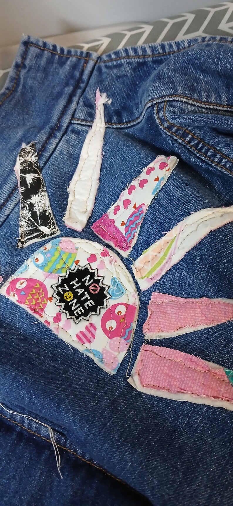 K&ouml;nnte beinhalten: Nahaufnahme eines Jeanskleidungsst&uuml;cks mit bunten Stoffflicken. Zu den Flicken geh&ouml;ren ein schwarz-wei&szlig;es Design, rosa Herzen und Eulen. Ein Aufn&auml;her lautet "NO HATE ZONE" in einem schwarzen Stern.
