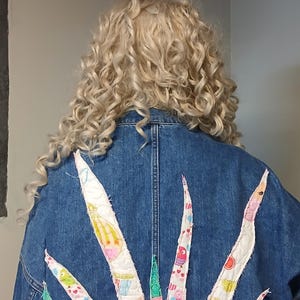 K&ouml;nnte beinhalten: Jeansjacke mit farbenfrohem Patchwork-Design auf dem R&uuml;cken. Das Design zeigt verschiedene Formen und Muster in Rosa-, Gr&uuml;n- und Wei&szlig;t&ouml;nen. Die Jacke hat einen ausgefransten Saum und wird von jemandem mit lockigem blondem Haar getragen.