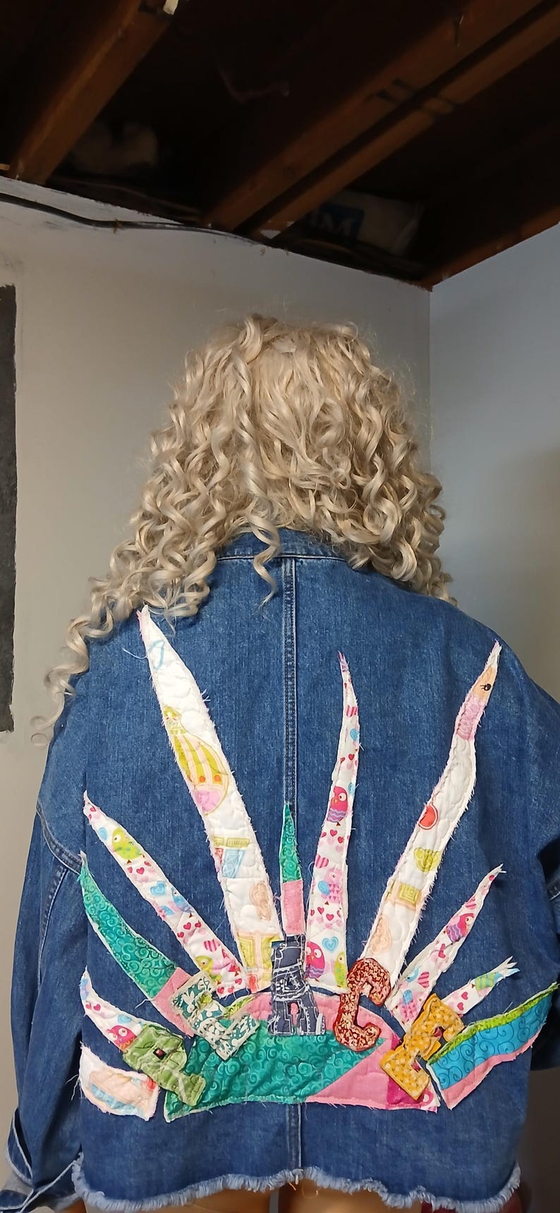 K&ouml;nnte beinhalten: Jeansjacke mit farbenfrohem Patchwork-Design auf dem R&uuml;cken. Das Design zeigt verschiedene Formen und Muster in Rosa-, Gr&uuml;n- und Wei&szlig;t&ouml;nen. Die Jacke hat einen ausgefransten Saum und wird von jemandem mit lockigem blondem Haar getragen.