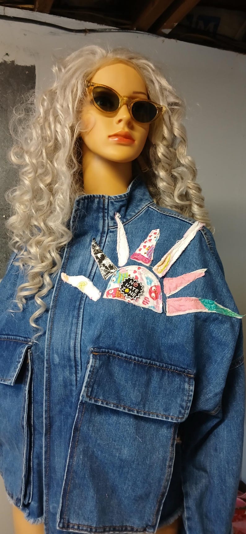 K&ouml;nnte beinhalten: Jeansjacke mit einem farbenfrohen Sonnenmuster auf der Brust, mit dem Text "NO HATE ZONE". Die Jacke hat zwei Vordertaschen und wird von einer Schaufensterpuppe mit Sonnenbrille getragen.