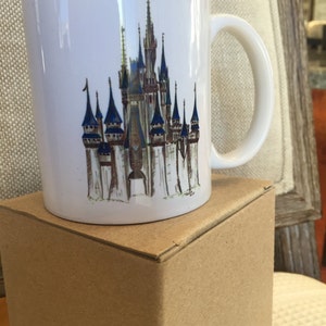 May include: A white ceramic mug with a colorful illustration of Cinderella's Castle from Walt Disney World. The mug has a handle and is sitting on a brown box.