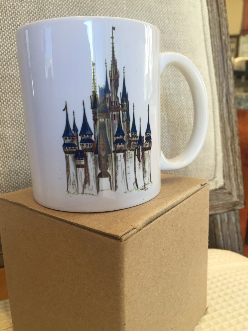 May include: A white ceramic mug with a colorful illustration of Cinderella's Castle from Walt Disney World. The mug has a handle and is sitting on a brown box.