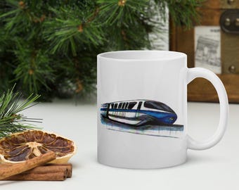 May include: A white ceramic mug featuring a detailed illustration of a blue and white monorail train. The train is depicted in a sleek, modern style with multiple windows and a streamlined shape, centered on the smooth surface of the mug.