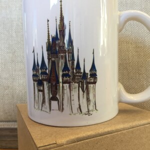 May include: White ceramic mug with a colorful illustration of Cinderella's Castle from Walt Disney World. The castle is depicted in a watercolor style with blue, gold, and brown accents.