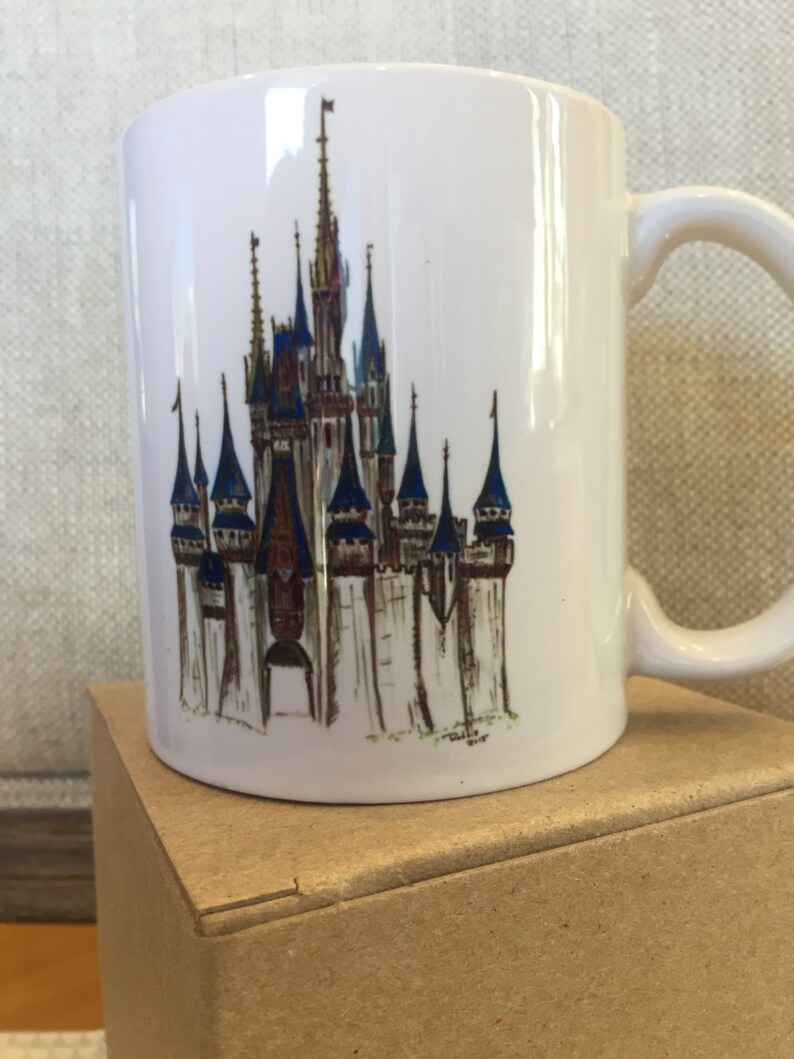May include: White ceramic mug with a colorful illustration of Cinderella's Castle from Walt Disney World. The castle is depicted in a watercolor style with blue, gold, and brown accents.