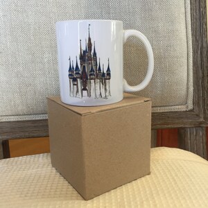 May include: White ceramic mug with a colorful illustration of Cinderella's castle on the front. The mug is sitting on a brown cardboard box.
