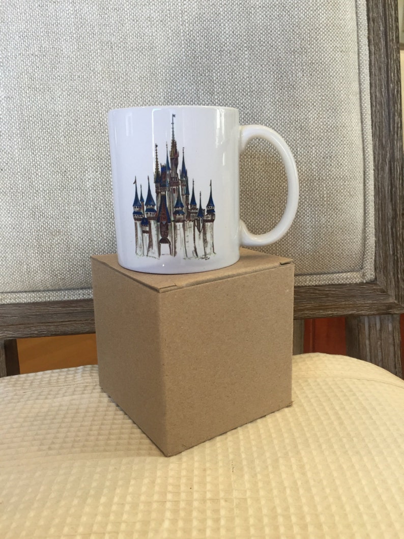 May include: White ceramic mug with a colorful illustration of Cinderella's castle on the front. The mug is sitting on a brown cardboard box.
