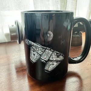 Może przedstawiać: Czarny ceramiczny kubek do kawy z białym obrazkiem statku kosmicznego Millennium Falcon z Gwiezdnych Wojen. Kubek ma małą, okrągłą, srebrną pokrywkę.