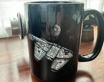 May include: Black ceramic coffee mug with a white image of the Millennium Falcon spaceship from Star Wars. The mug has a small, round, silver lid.