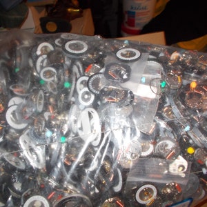 May include: A clear plastic bag filled with numerous watch components. The components include watch faces, dials, and other small parts, with a mix of silver, black, and white colors. Some parts have small, colorful accents.