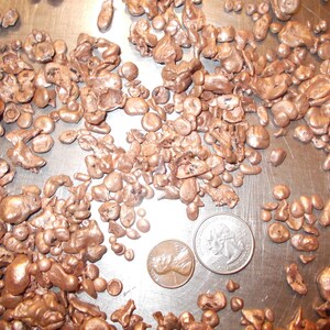 May include: A close-up of a pile of copper nuggets on a silver surface. The nuggets are various sizes and shapes, with some being round and others being more irregular. Two US coins are included for scale.
