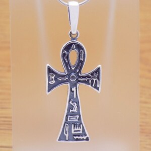 May include: A silver-toned ankh pendant with black detailing and hieroglyphic symbols. The pendant is suspended from a thin silver chain. The ankh is a symbol of life and immortality in ancient Egyptian culture.