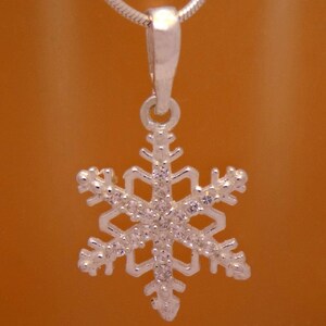 May include: A silver snowflake pendant with clear crystals. The pendant is suspended from a silver chain. The snowflake design features six symmetrical points, each adorned with small, sparkling crystals. The background is a warm, orange-brown color.