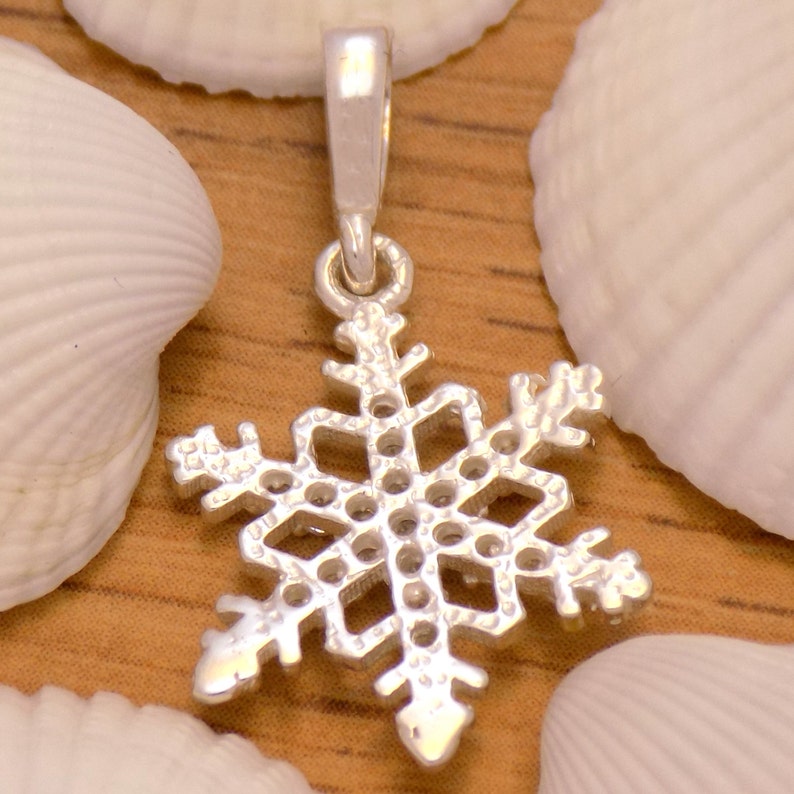 May include: A silver snowflake pendant with a loop for a chain. The pendant is detailed with cut-out designs and is set against a background of seashells and a wooden surface. The snowflake design is a winter theme.