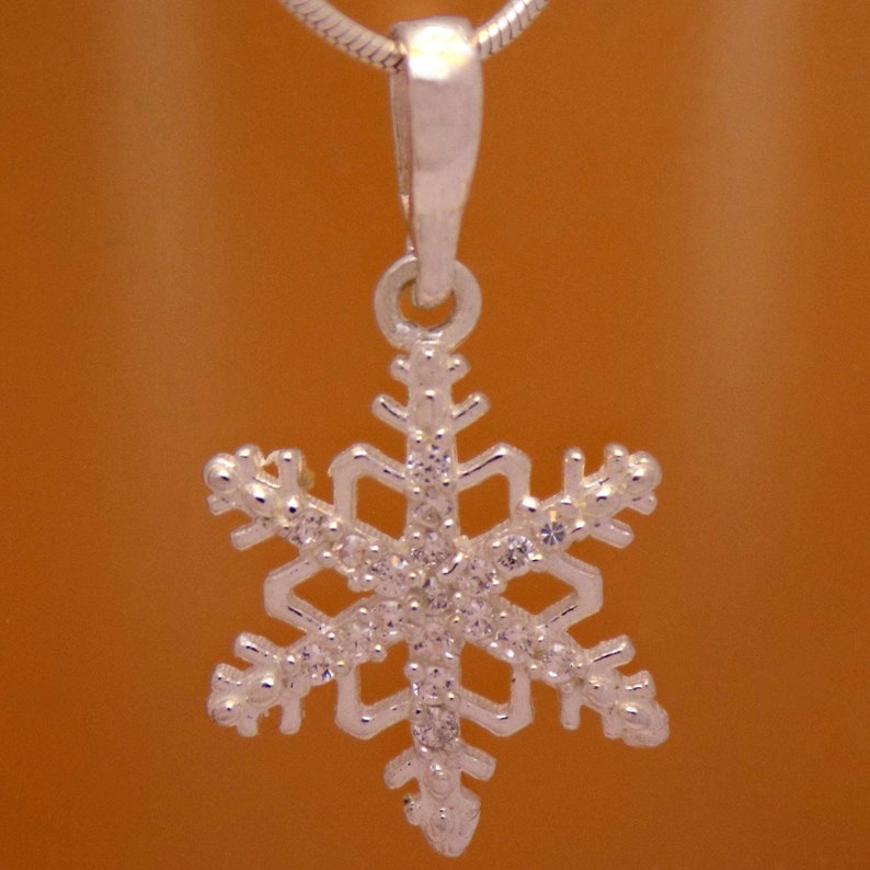 May include: A silver snowflake pendant with clear crystals. The pendant is suspended from a silver chain. The snowflake design features six symmetrical points, each adorned with small, sparkling crystals. The background is a warm, orange-brown color.